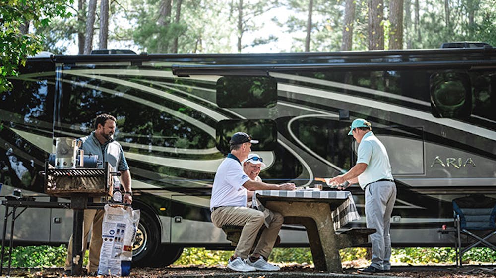 2021 Thor Aria Class A Diesel Pusher RV Lifestyle side view men grilling and eating at campsite