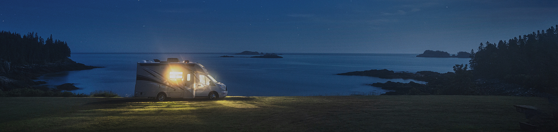A motor home parked next to the shore at night with the light on illuminating its surroundings